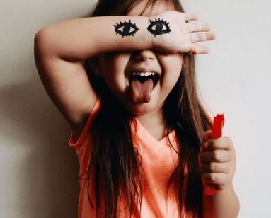 A young girl covers her eyes with her arm and sticks out her tongue. Two eyes are drawn on her arm over her own eyes.
